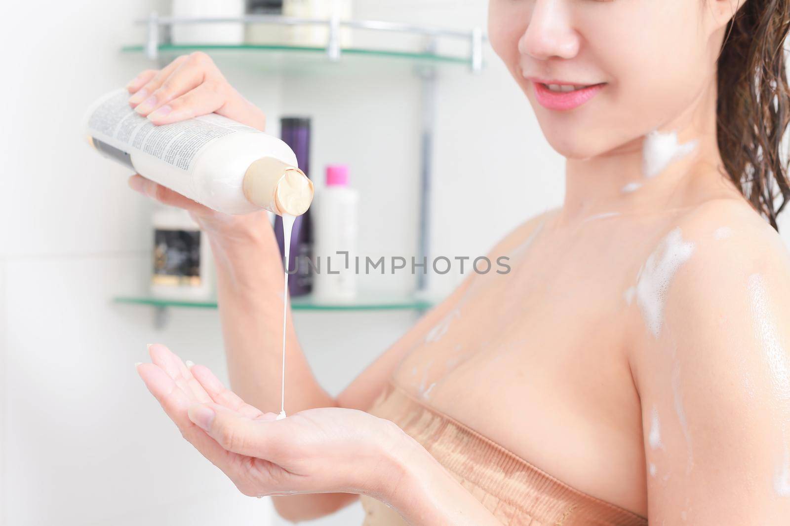 Woman in bath towel is applying cream on her hand while standing in front of the mirror in bathroom.