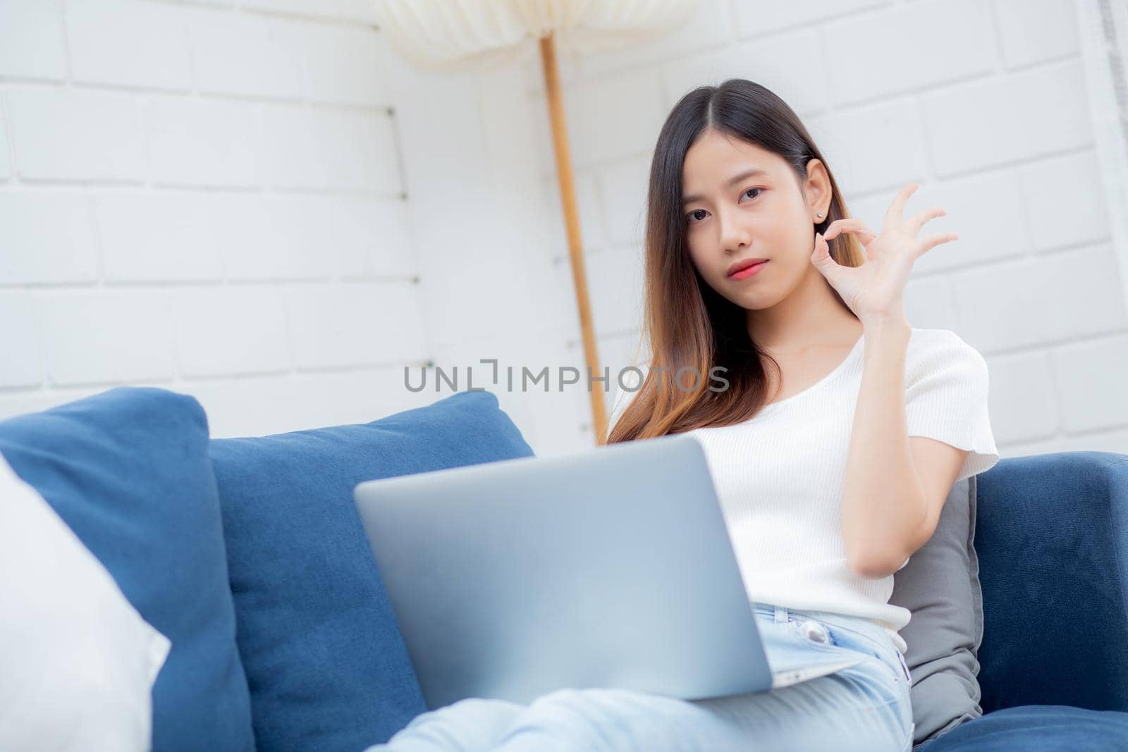 Young asian business woman smile and work from home with laptop computer online to internet on sofa in living room, freelance girl using notebook on couch with comfort, new normal, lifestyle concept. by nnudoo