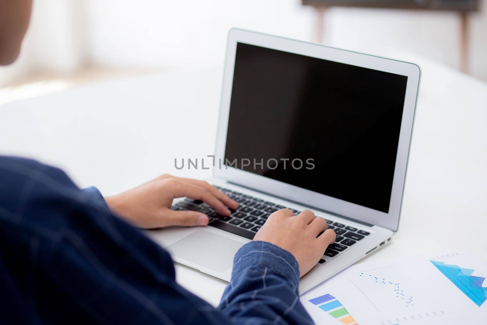 Young asian business man working laptop computer blank display screen while report graph and chart paper on desk, businessman analysis statistic of finance on table, planning for success, indoor. by nnudoo