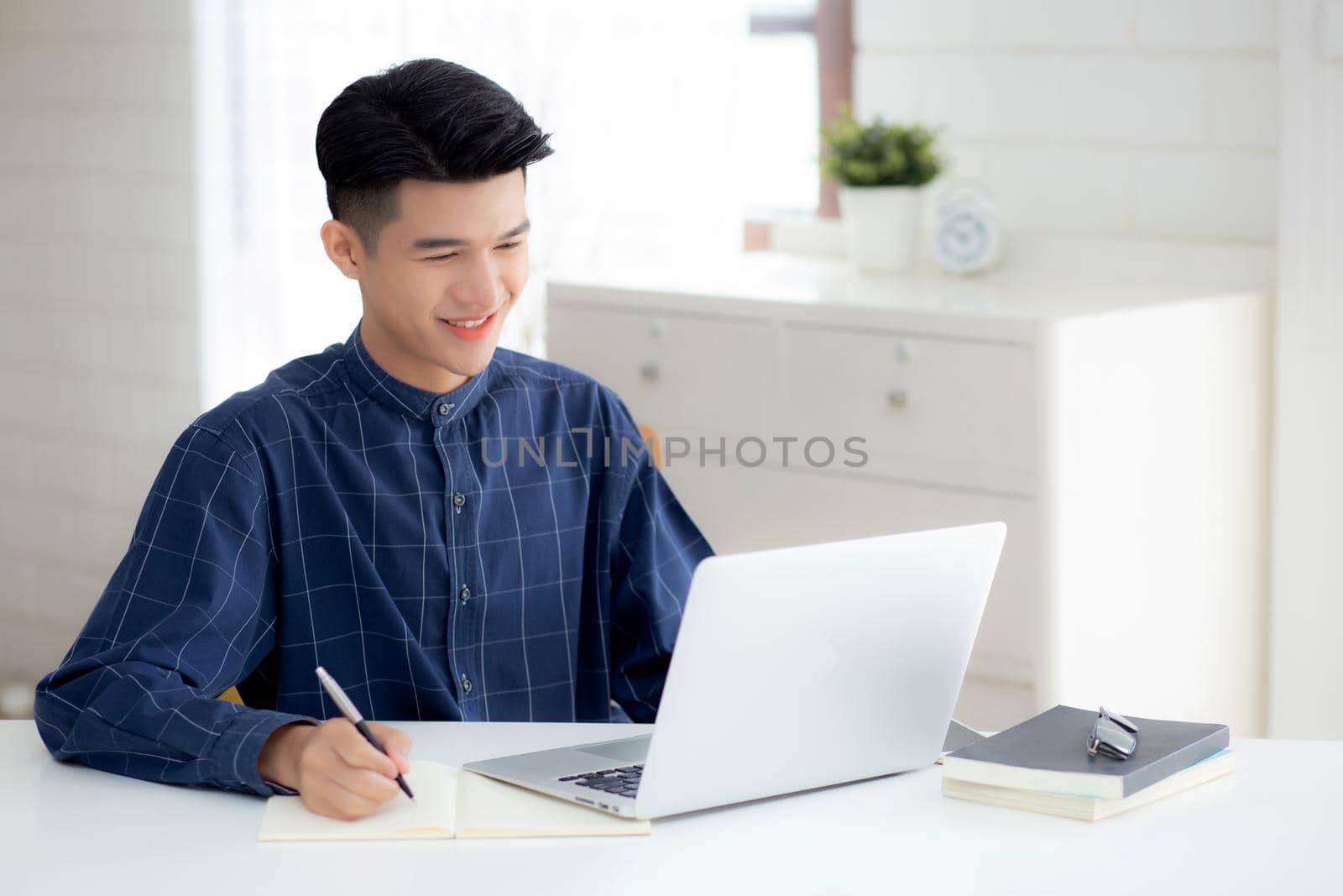 Young asian business man writing on notebook for planning working and using laptop computer on desk at home, notes about finance, male study and learning, business and communication concept. by nnudoo