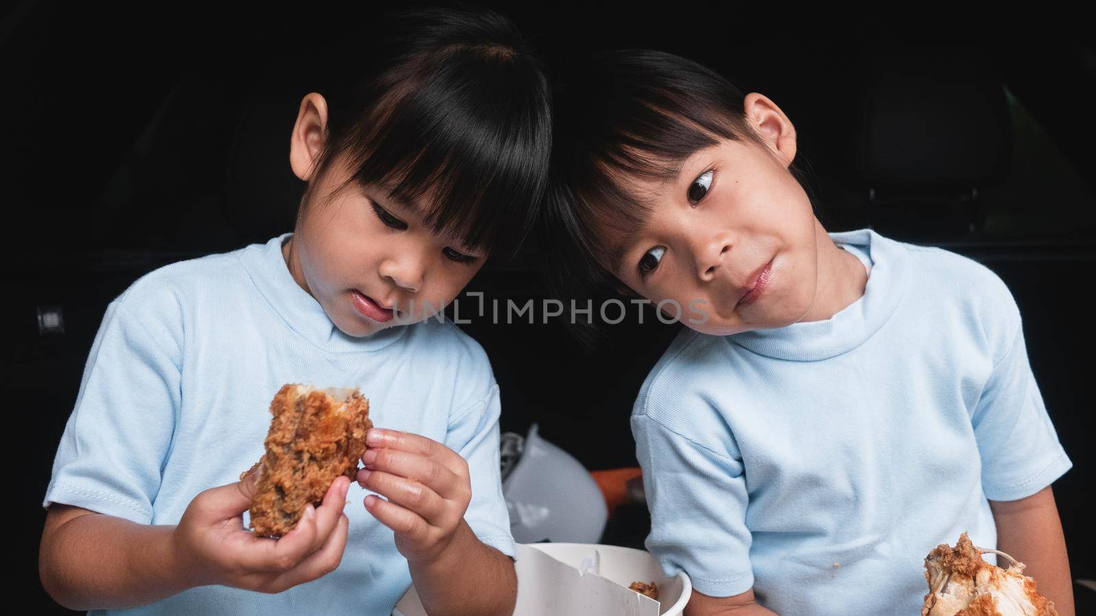 Two sibling girls having break during family road trip and eating fried chicken in trunk of car on the park. Delivery service and Social distance from Covid-19 or Coronavirus.