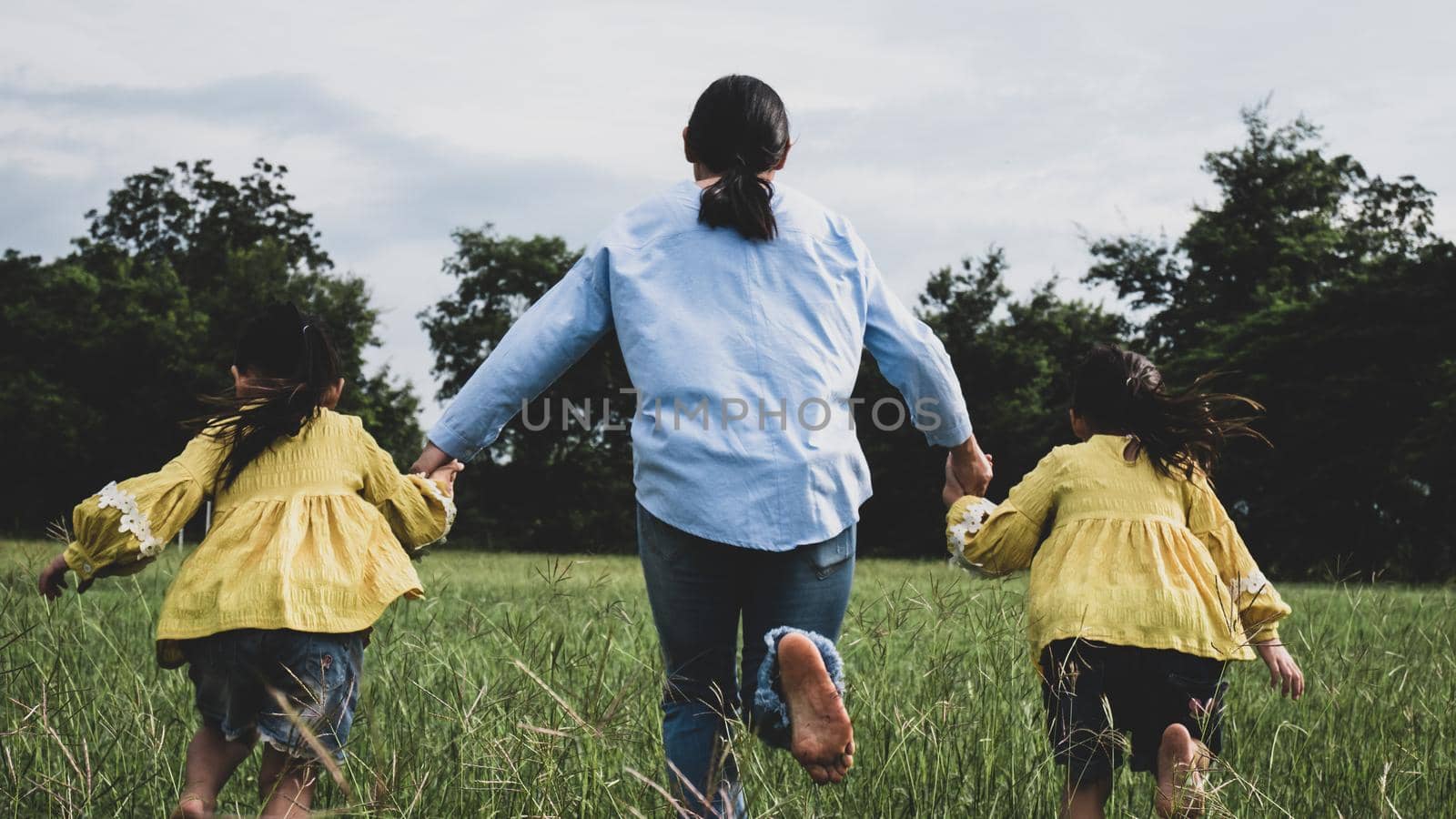 Mother and daughters holding hands and running on field in summer park. Happy family spend their free time together on vacation. by TEERASAK