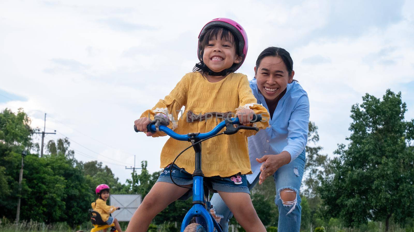 Cute little girl in safety helmet learn to ride a bike with her mother in summer park. Outdoor sports for kids. childhood happiness. family spending time together. by TEERASAK