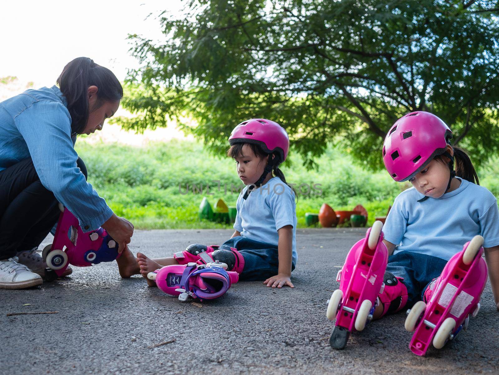 Young mother helps her daughter put on protective pads and a safety helmet before her roller skating practice on the park road. Active outdoor sport for kids.