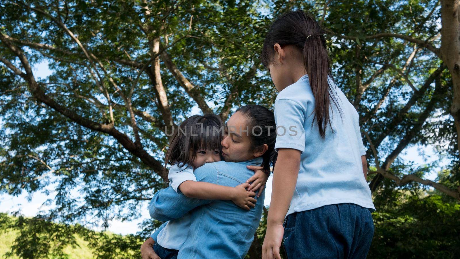 Two sibling little girls with mom laughing and hugging each other on warm and sunny summer day in the garden. Young girls with her mother spending day in park.