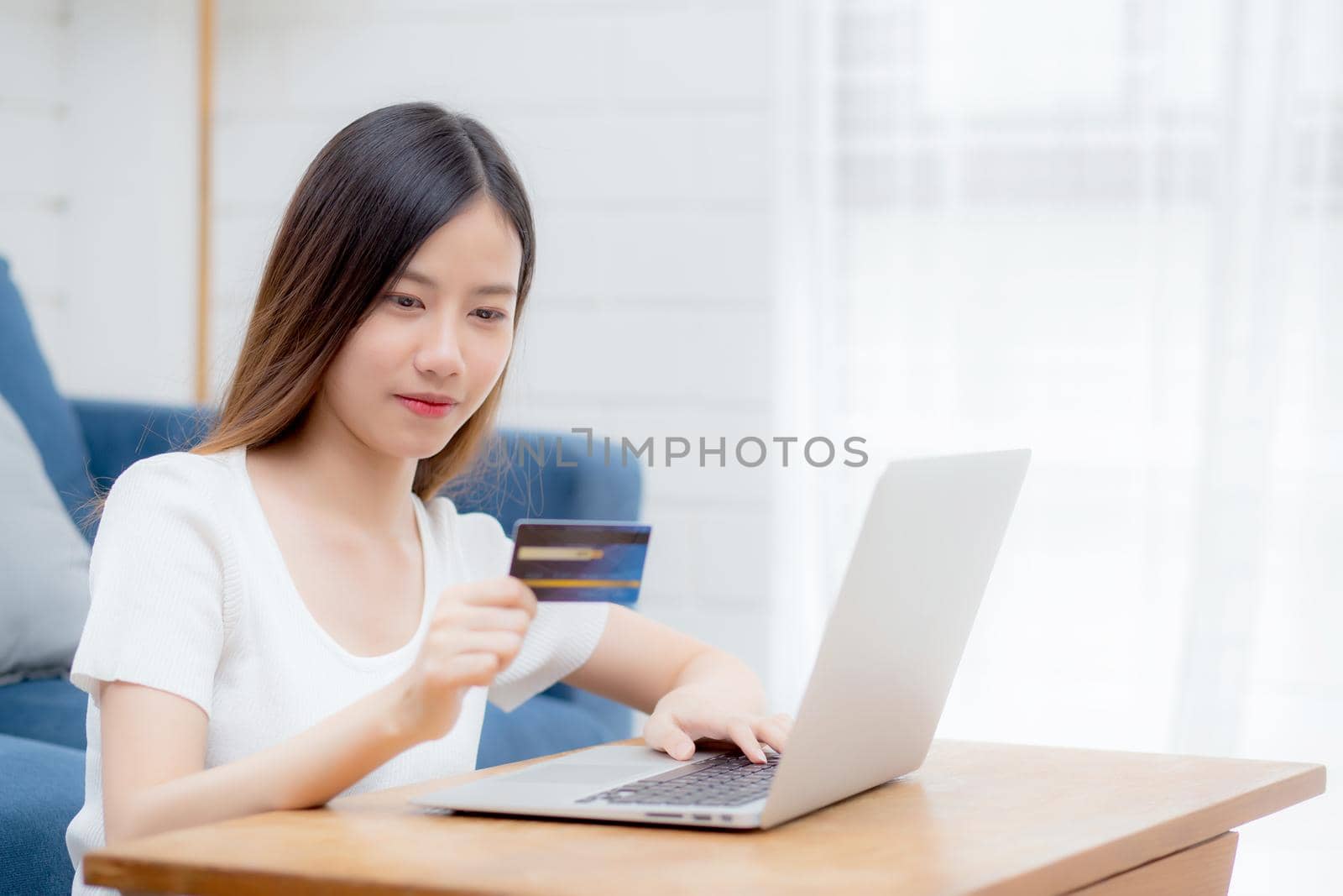 Young asian woman sit on sofa using laptop computer shopping online with credit card buying to internet, happy girl payment with e-business on couch, purchase and payment, business concept. by nnudoo