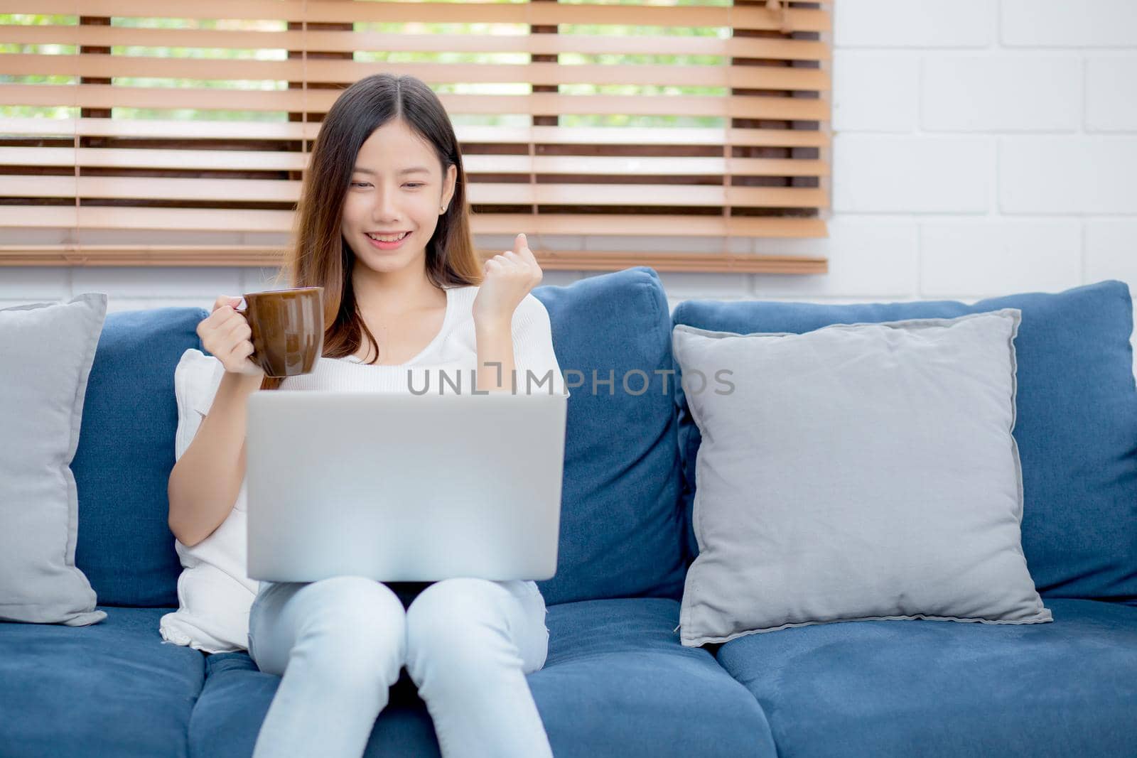 Young asian business woman smile and work from home with laptop computer online on sofa in living room, freelance girl using notebook and drink coffee on couch, new normal, lifestyle concept.