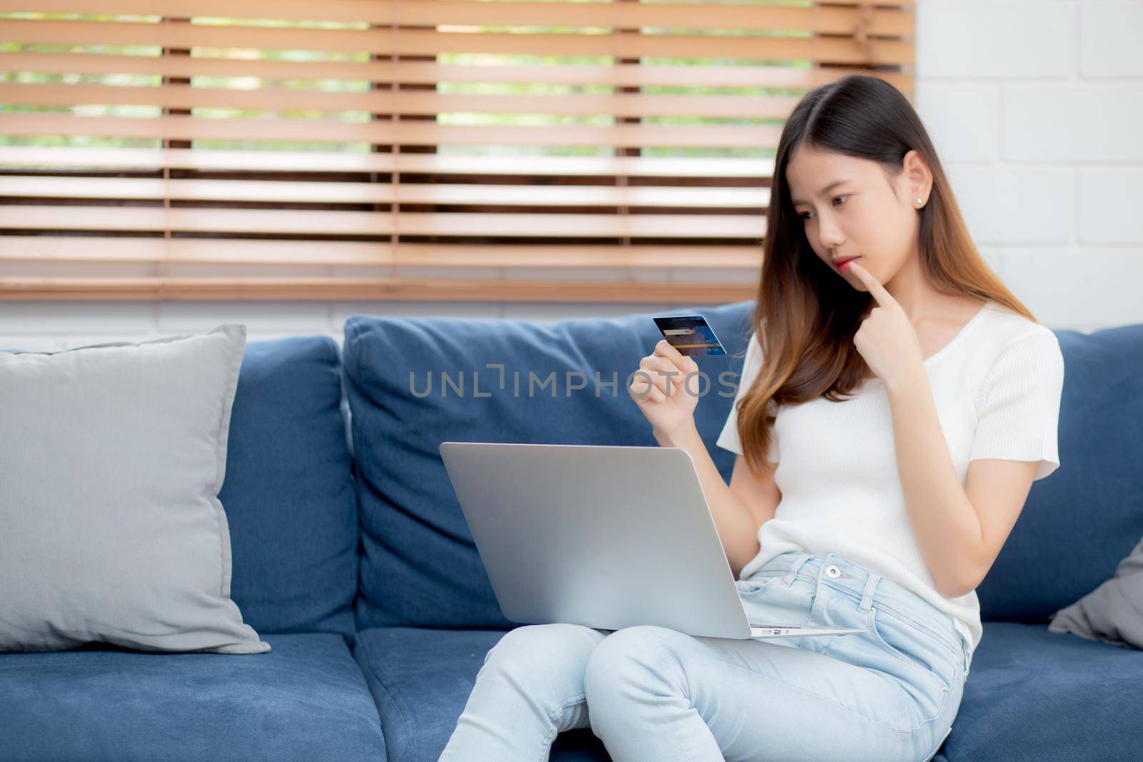 Young asian woman sitting thinking idea using credit card with laptop computer on couch, girl shopping online for buy and payment with notebook on sofa, finance and debit, lifestyle concept.