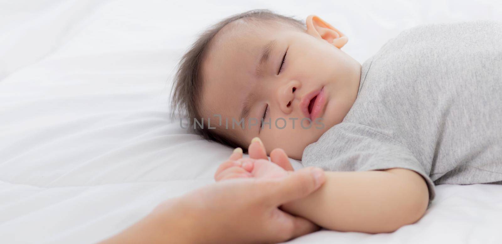 Young asian mother touch hand of little baby girl with tender on bed in the bedroom, mom love newborn and care, woman with expression with child together, parent and daughter, family concept.