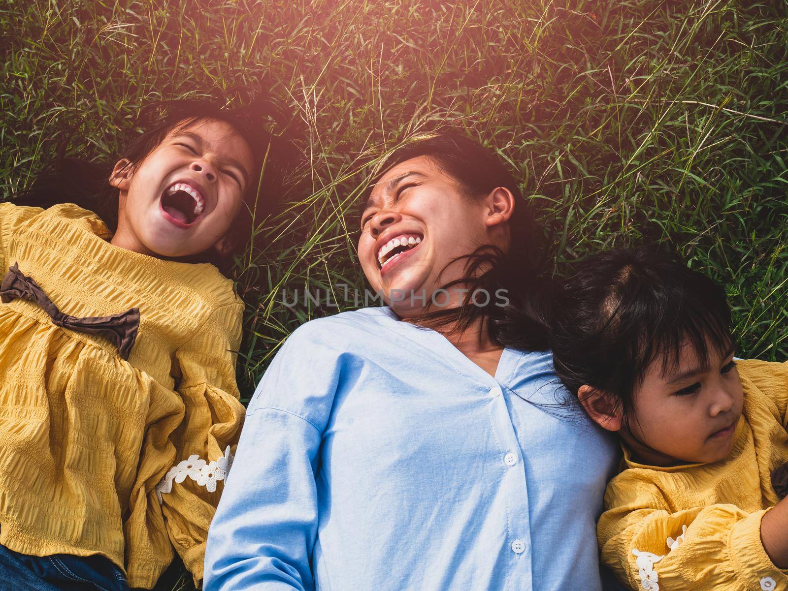 Happy mother and daughters smiling and laughing lying on the lawn in summer park. Family spend their free time together on vacation. by TEERASAK