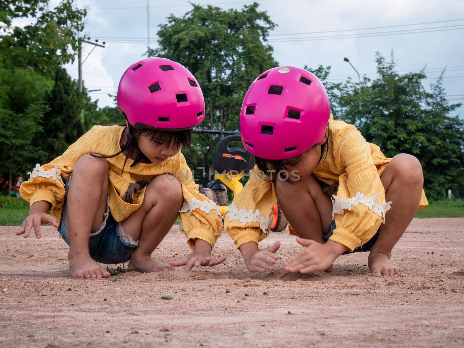 Two cute little sisters in helmets are playing with sand in park on a sunny summer day, taking a break after cycling practice. develop imagination and exploration. by TEERASAK