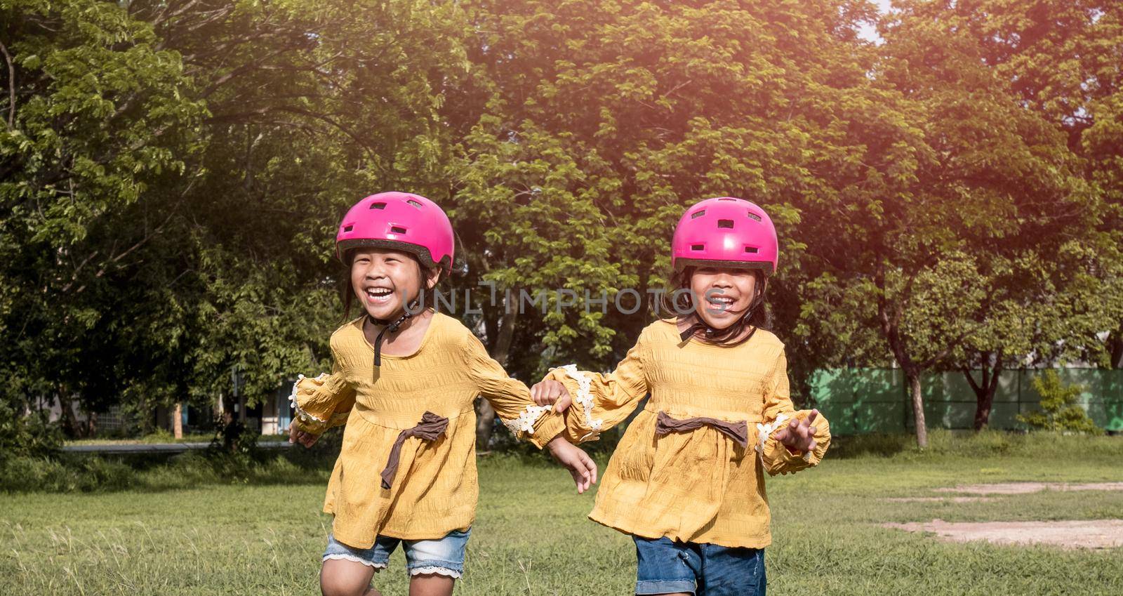 Two cute little sisters in helmet running on the lawn in park on a sunny summer day, taking a break after cycling practice. Happy childhood. Family spending time together. by TEERASAK