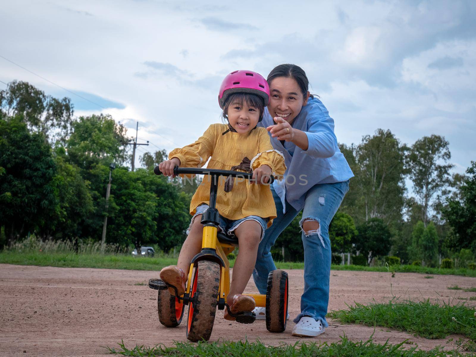 Cute little girl in safety helmet learn to ride a bike with her mother in summer park. Outdoor sports for kids. childhood happiness. family spending time together. by TEERASAK