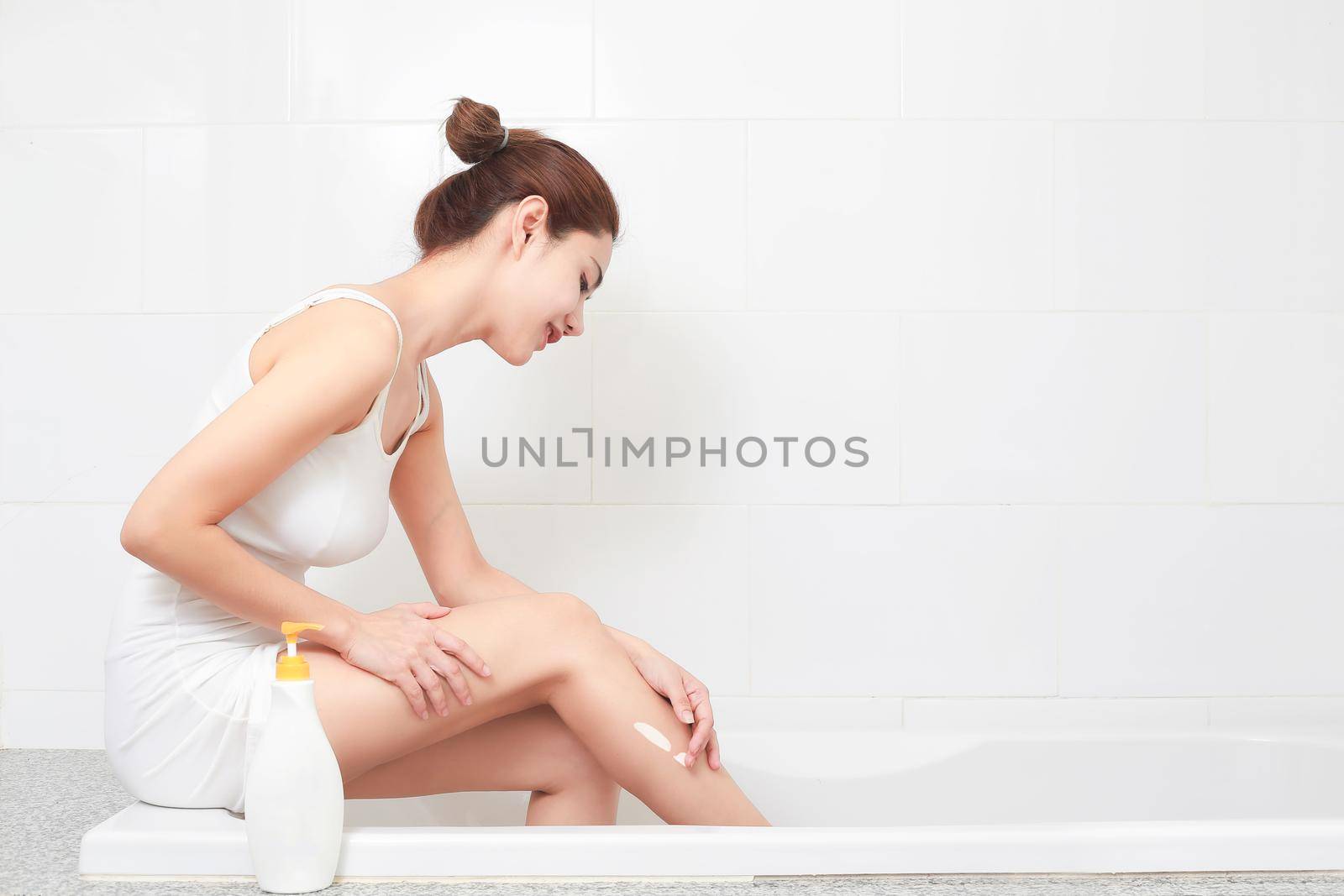 Young beautiful woman applying body lotion on her attractive legs in bathroom.