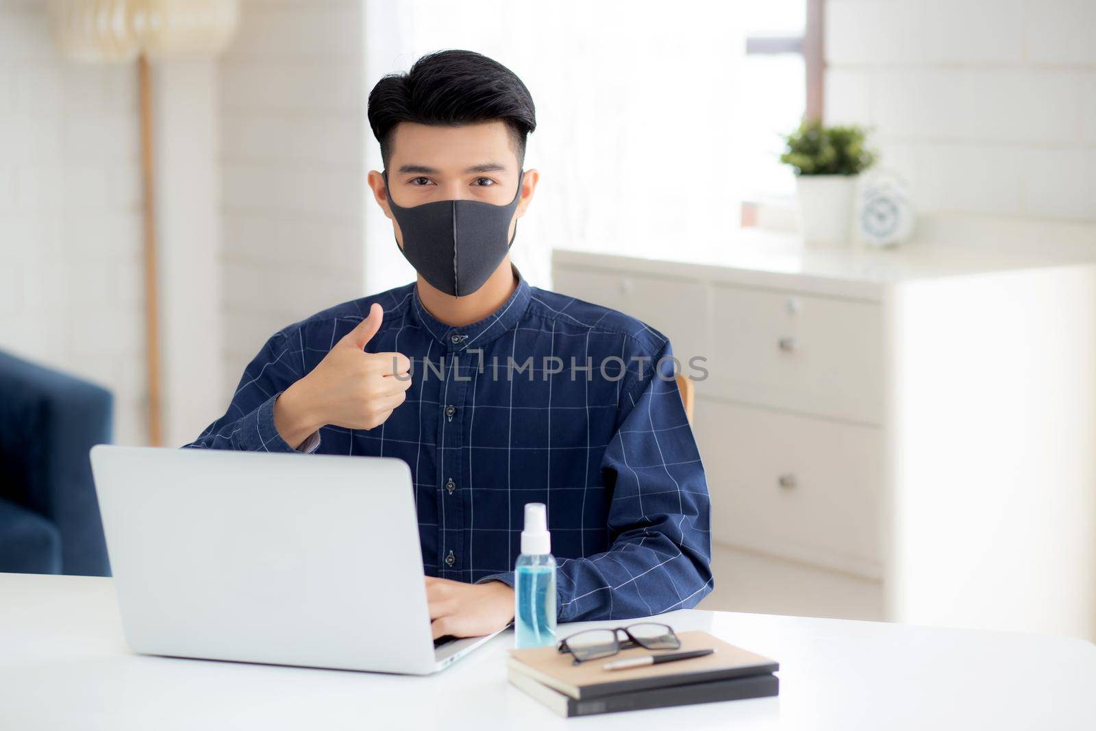 Young business man in face mask working from home with laptop computer on desk for protection for outbreak of covid-19 and healthcare, male quarantine stay home using notebook on desk, new normal. by nnudoo