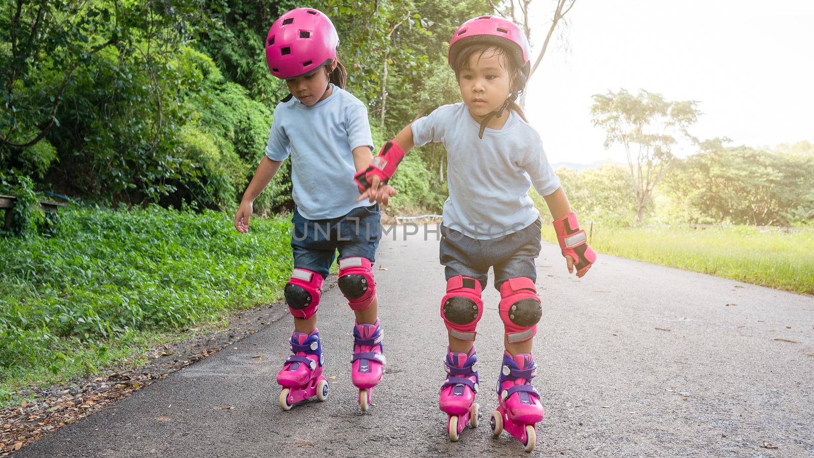 Two sibling sisters wearing protection pads and safety helmet practicing to roller skate on the street in the park. Active outdoor sport for kids. by TEERASAK