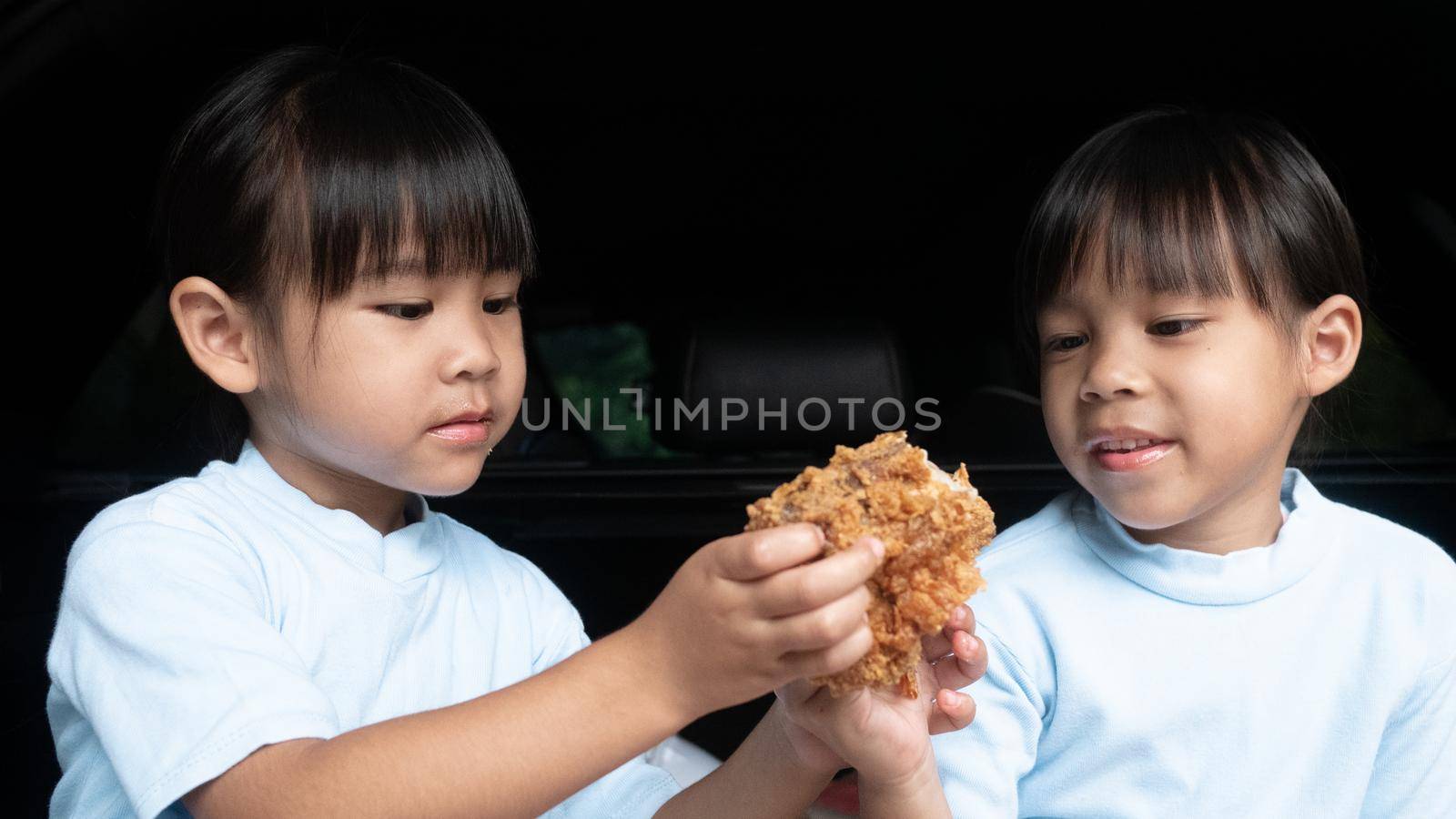 Two sibling girls having break during family road trip and eating fried chicken in trunk of car on the park. Delivery service and Social distance from Covid-19 or Coronavirus. by TEERASAK