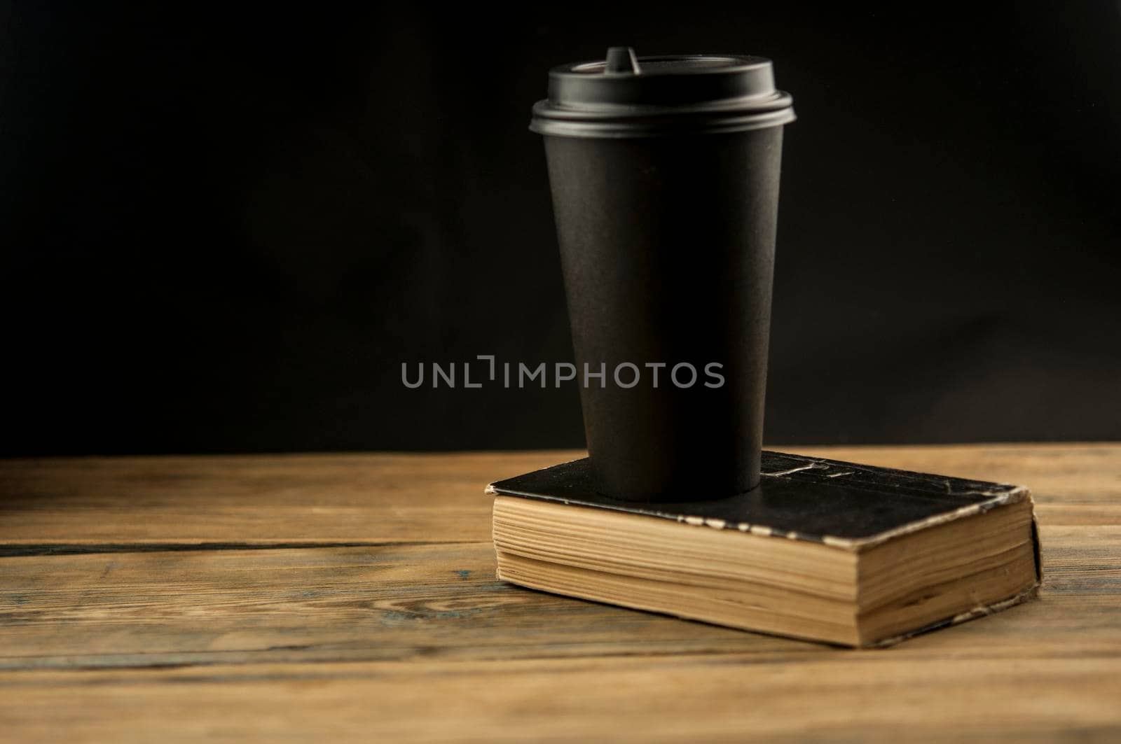Old book and a cup of coffee in a disposable paper cup on a wooden table by inxti