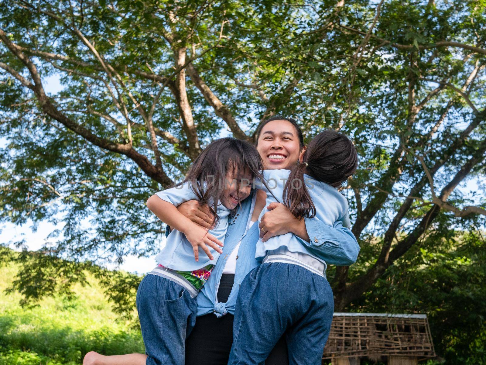 Two sibling little girls with mom laughing and hugging each other on warm and sunny summer day in the garden. Young girls with her mother spending day in park. by TEERASAK