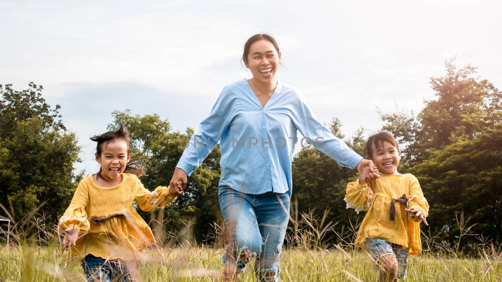 Mother and daughters holding hands and running on field in summer park. Happy family spend their free time together on vacation. by TEERASAK