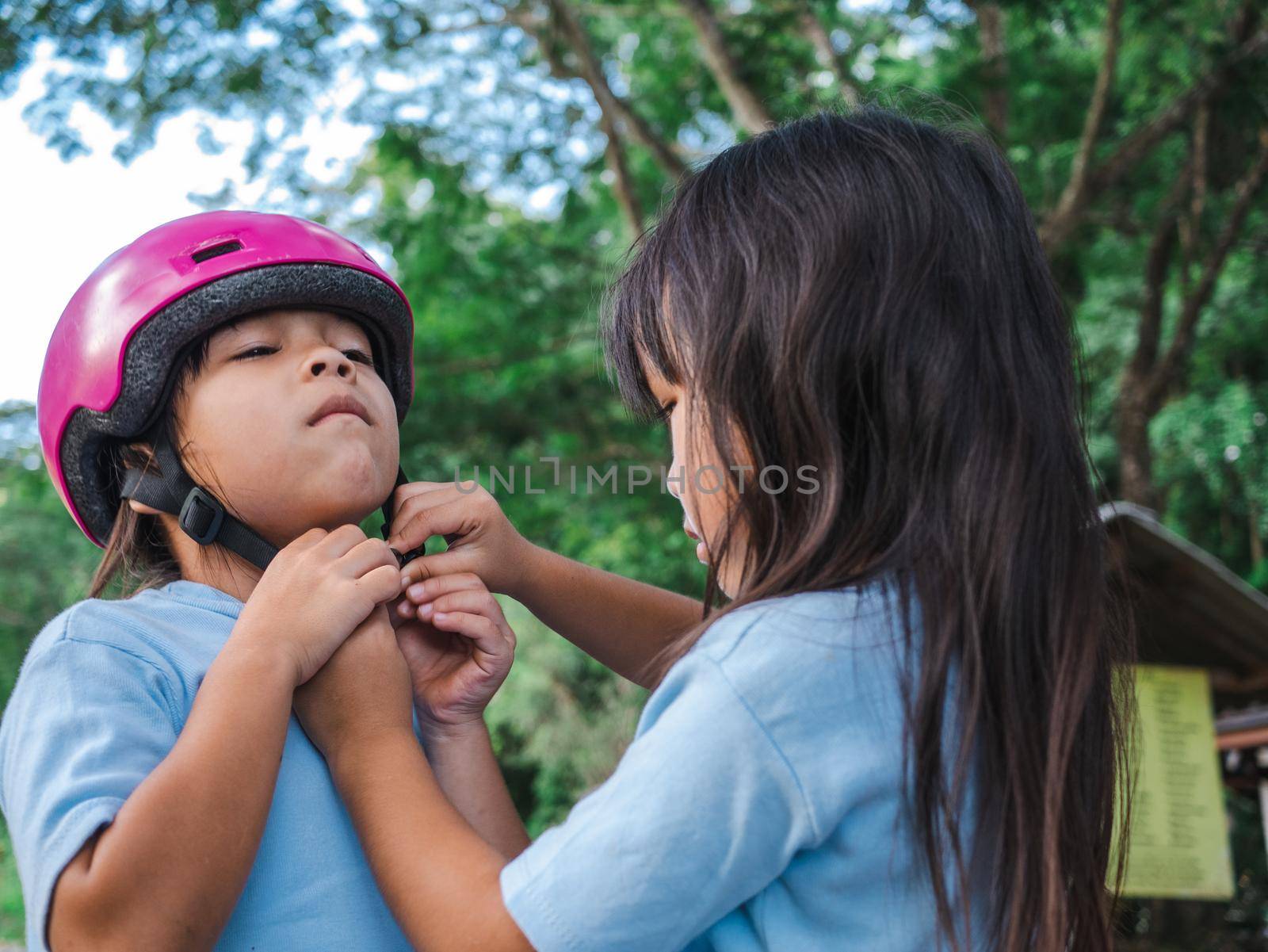 Cute older sister is helping younger sister to put on a protection pads and safety helmet practicing to roller skate on the street in the park. Active outdoor sport for kids. by TEERASAK