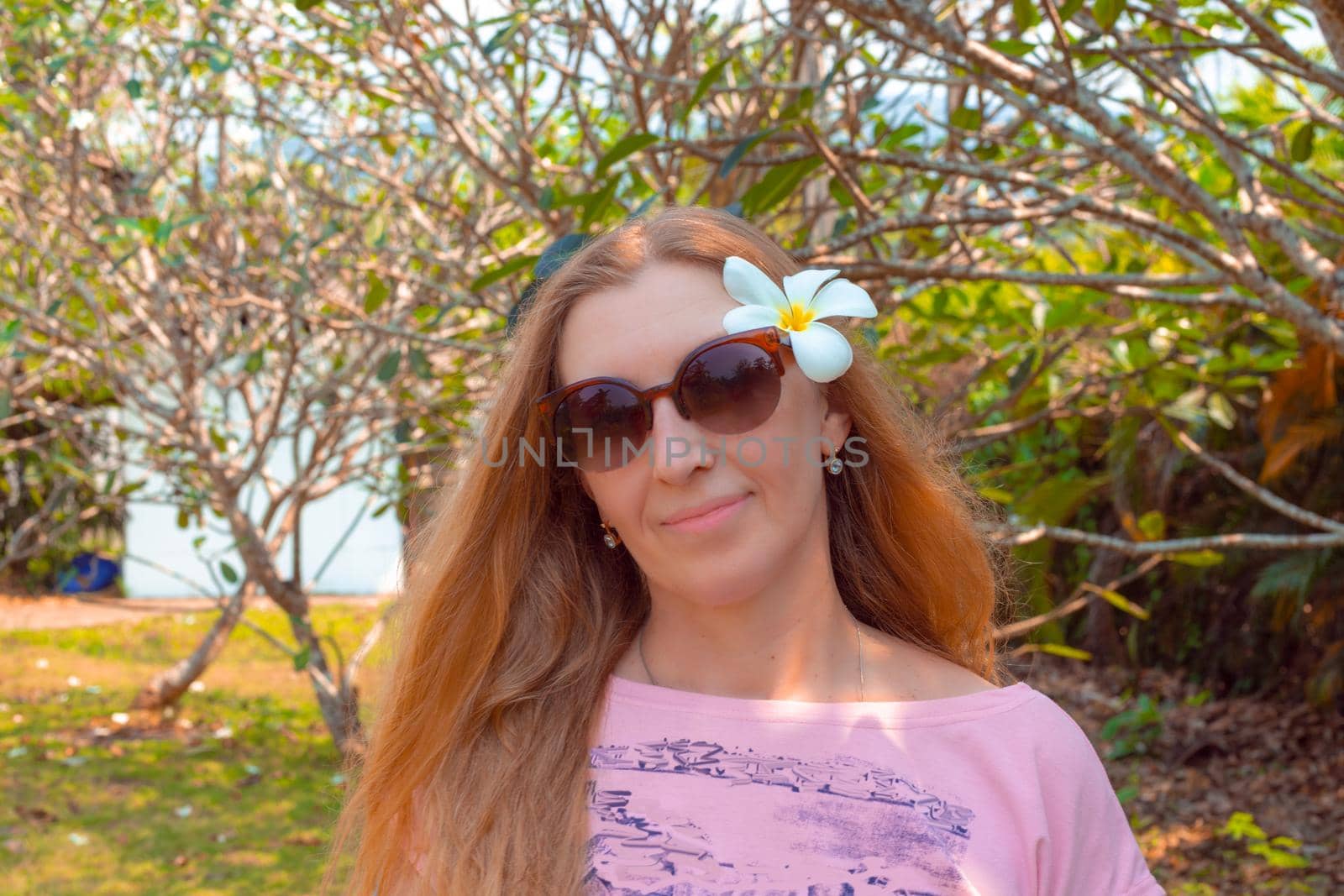 girl smiles with a white plumeria flower in blond long hair, in sunglasses, in the tropics