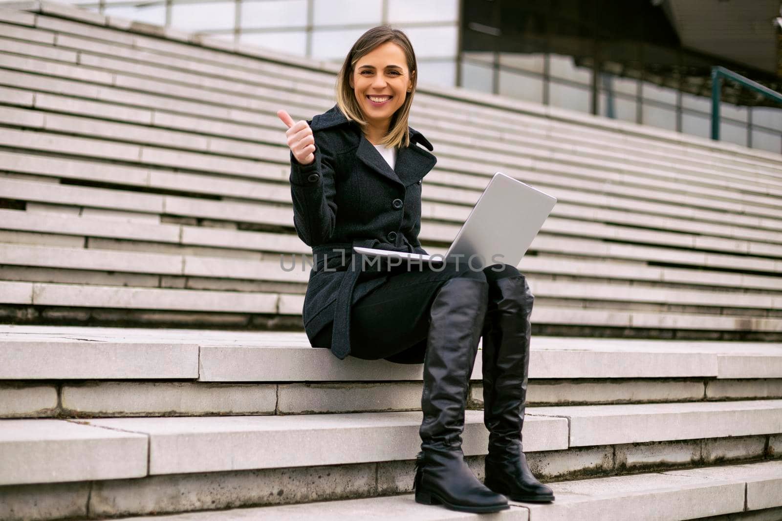 Beautiful businesswoman using laptop and showing thumb up while sitting on the staircase in the city.