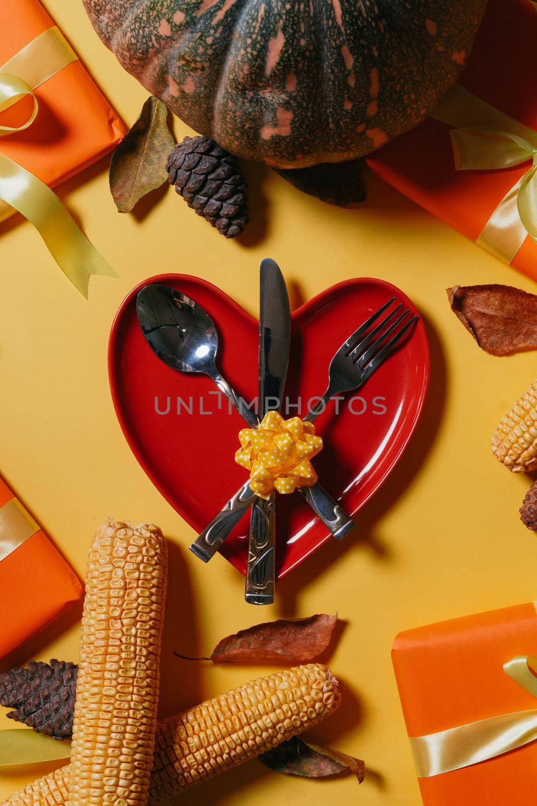 Happy thanksgiving day. Heart shape plate, fork, spoon and knife with harvest fall vegetables and autumnal leaves on yellow background. Autumnal background