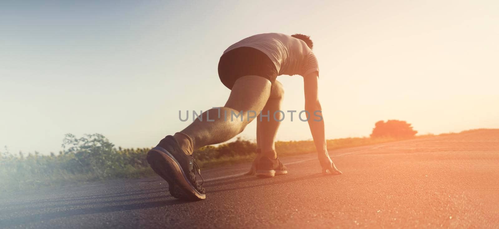 A young man prepares for a quick jog on an asphalt road, around the field. A runner warms up, stretches before training at sunset, legs close-up.