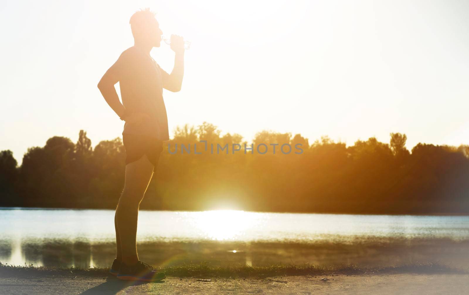 A young man drinks water while jogging along the embankment near the lake in the park. Athlete runner training early in the morning in nature.