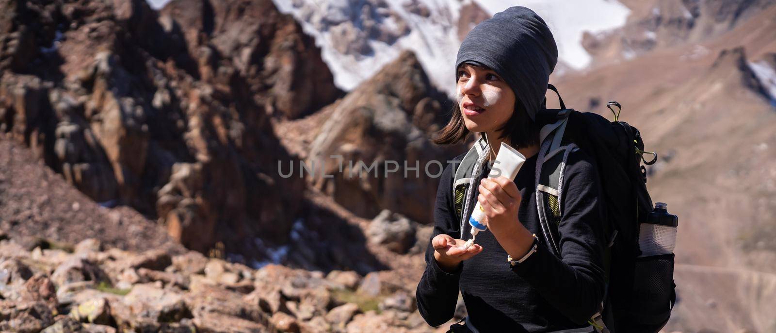 Beautiful young sportive girl applies sunscreen to her hands and face while climbing the mountain, woman takes care of her skin while traveling.