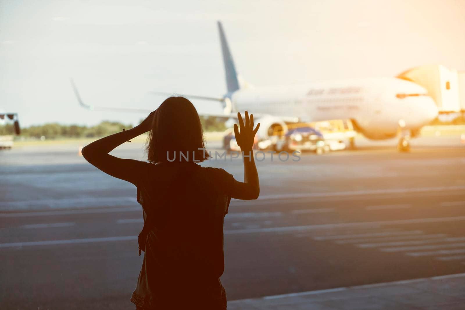 A young girl at the airport waving to the plane on the runway at sunset. Silhouette of a traveler near the window in the transfer area.