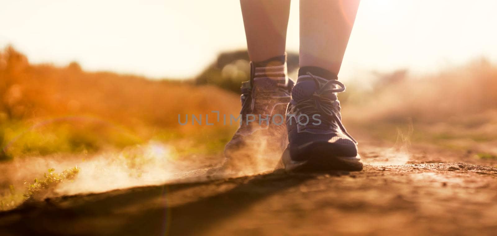 Female athletic legs in sneakers at sunset during outdoor jogging at sunset. A young girl warms up, she is engaged in trail running, view on her legs and clouds of dust close-up.