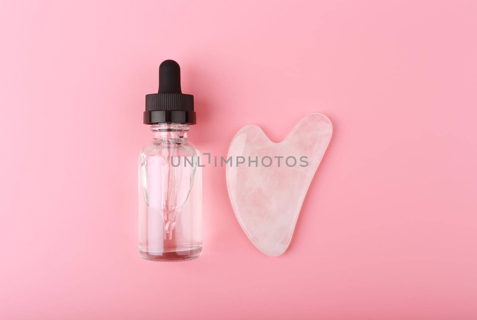 Flat lay with massage gel, skin serum or oil in transparent glass bottle and heart shaped guasha pink quartz crystal on bright pink background. Concept of facial massage and self treatment