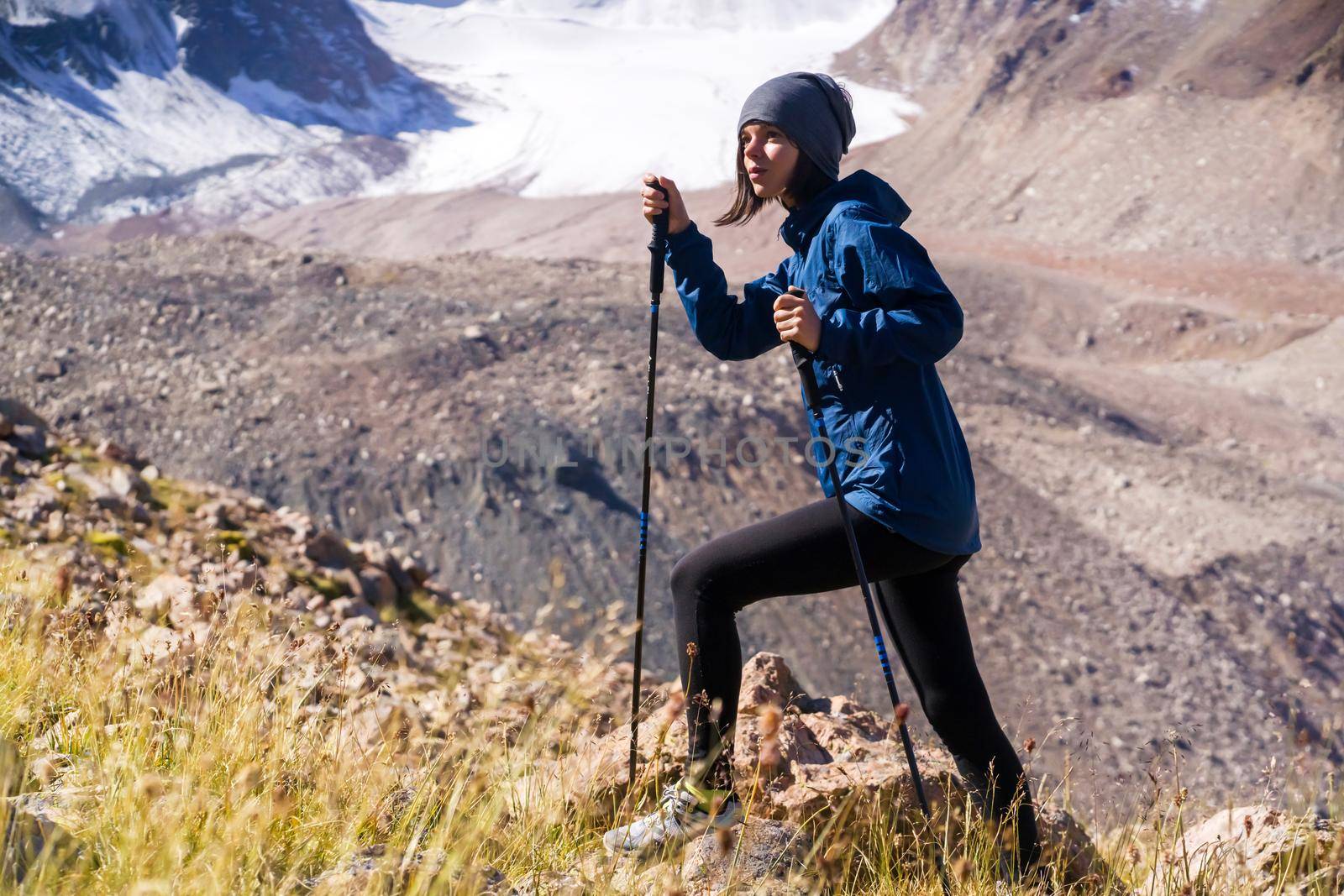 A young girl leads an active lifestyle, climbs a trail in the mountains with trekking poles, snow-capped mountain peaks and a glacier in the background.