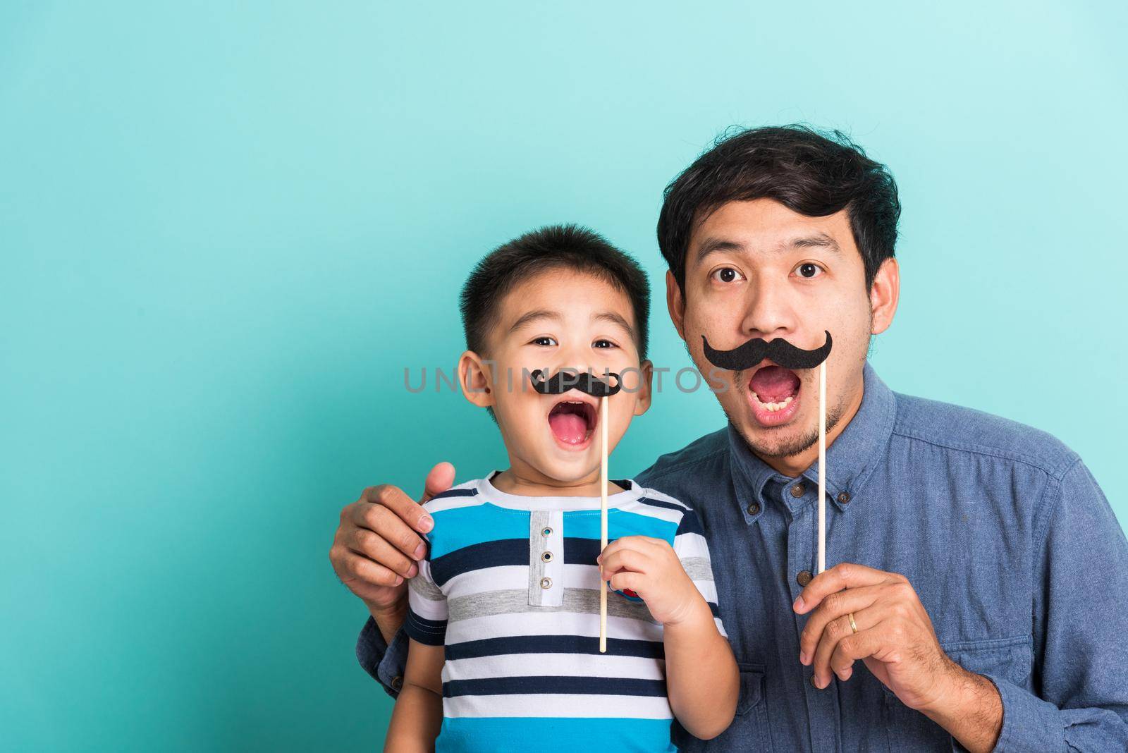 Family funny happy hipster father and his son kid holding black mustache props for the photo booth close face, studio shot isolated on a blue background, November men health awareness