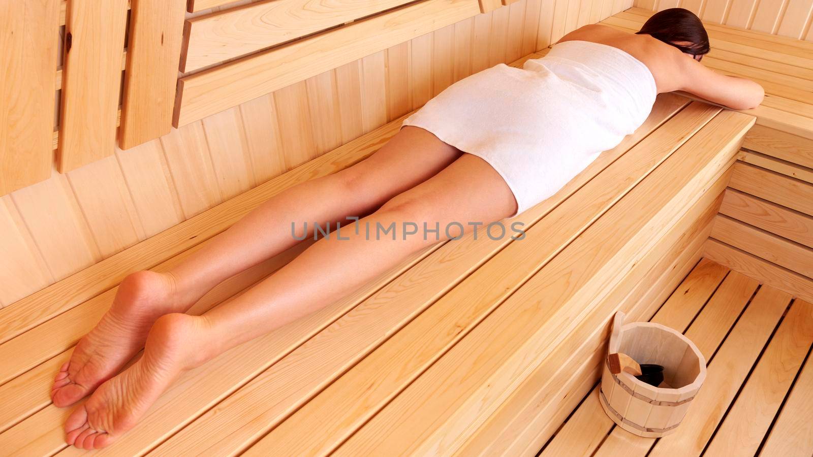 Woman relaxing in a wooden sauna lying on her belly on the bench with a towel wrapped around her body by Nobilior