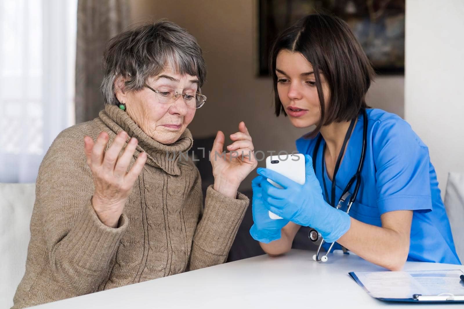 A nurse in a medical suit takes care and explains to an elderly patient how to use applications on a smartphone. Grandmother 80 years old, does not understand how to communicate with a doctor online.