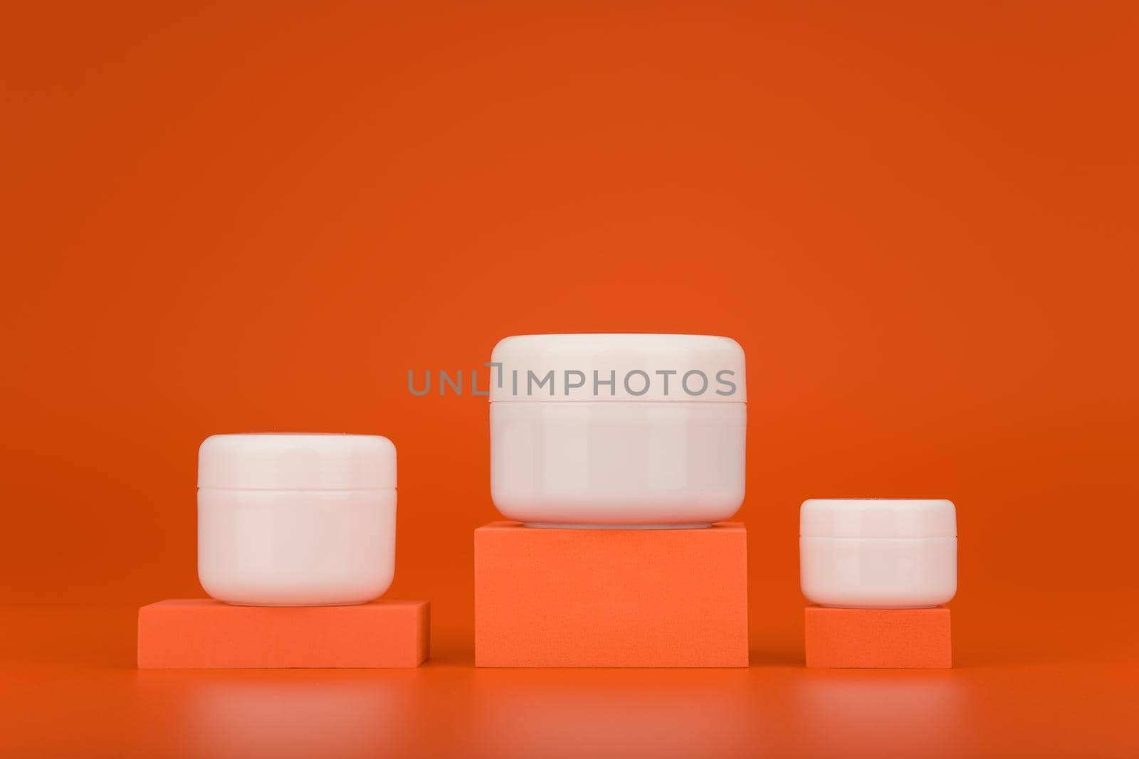 Set of three cosmetic jars of different sizes on podiums against orange background with copy space. Concept of cream, lotion, mask or scrub with vitamin C for anti aging or regular skin care