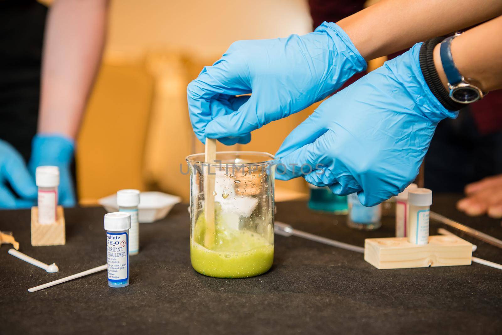 Up close view of young scientist wearing blue latex gloves stirring green liquid in a chemistry glass with surrounding vials for testing.