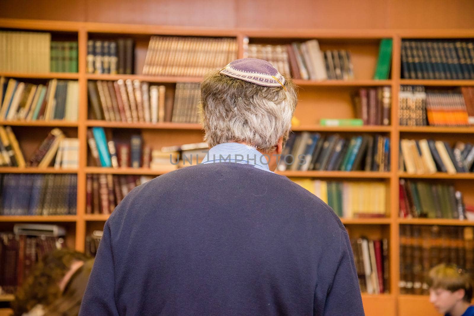 Old Jewish man with gray hair wearing a yarmulke from the back in a library of book shelves.