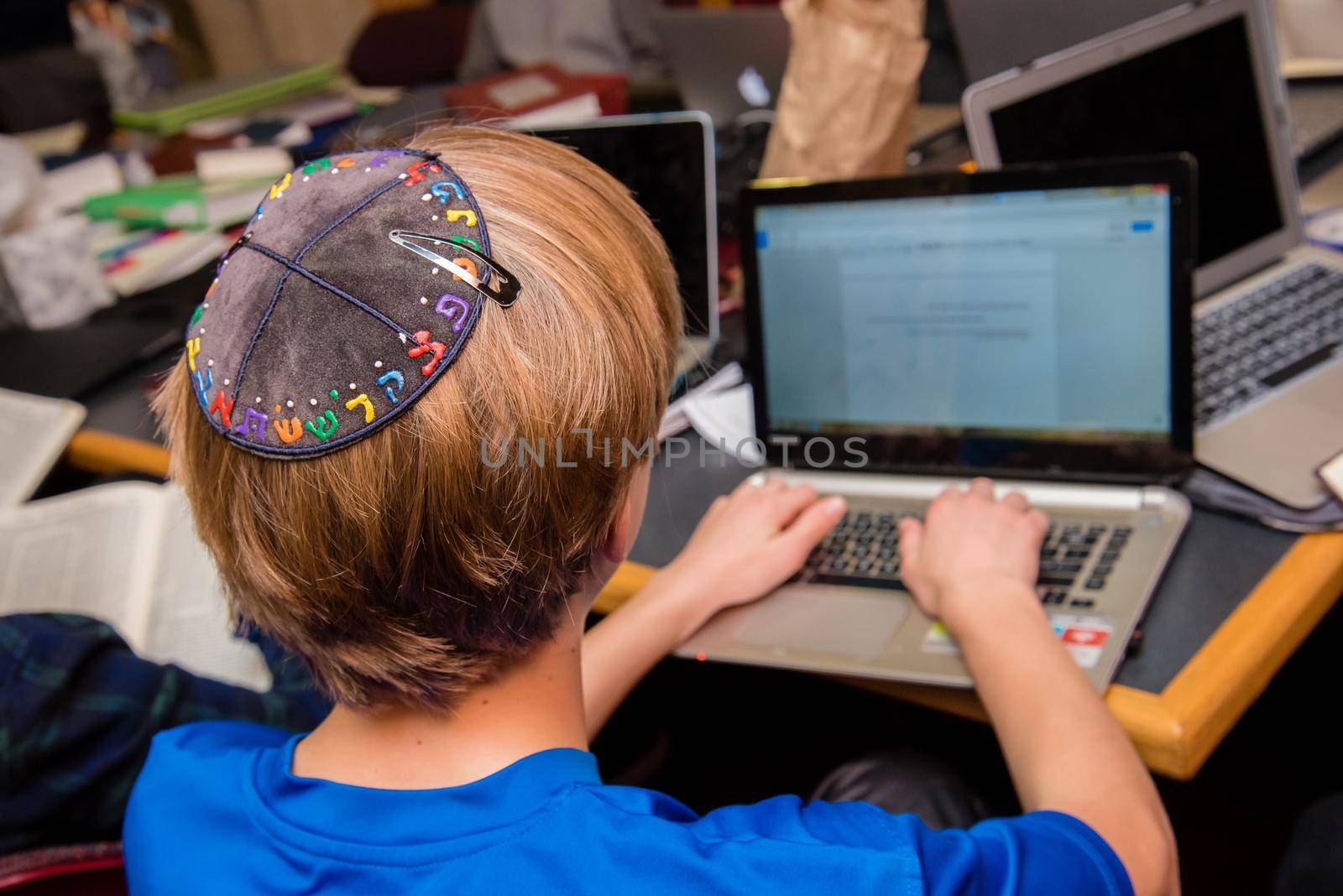Jewish boy wearing yarmulke from the back sitting in a classroom setting with students. by jyurinko