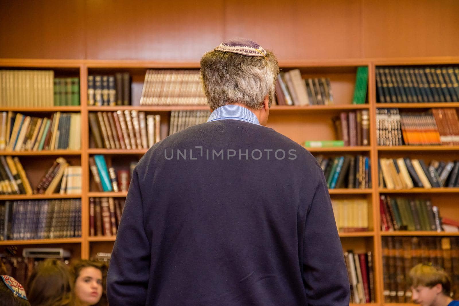 Old Jewish man with gray hair wearing a yarmulke from the back in a library of book shelves.
