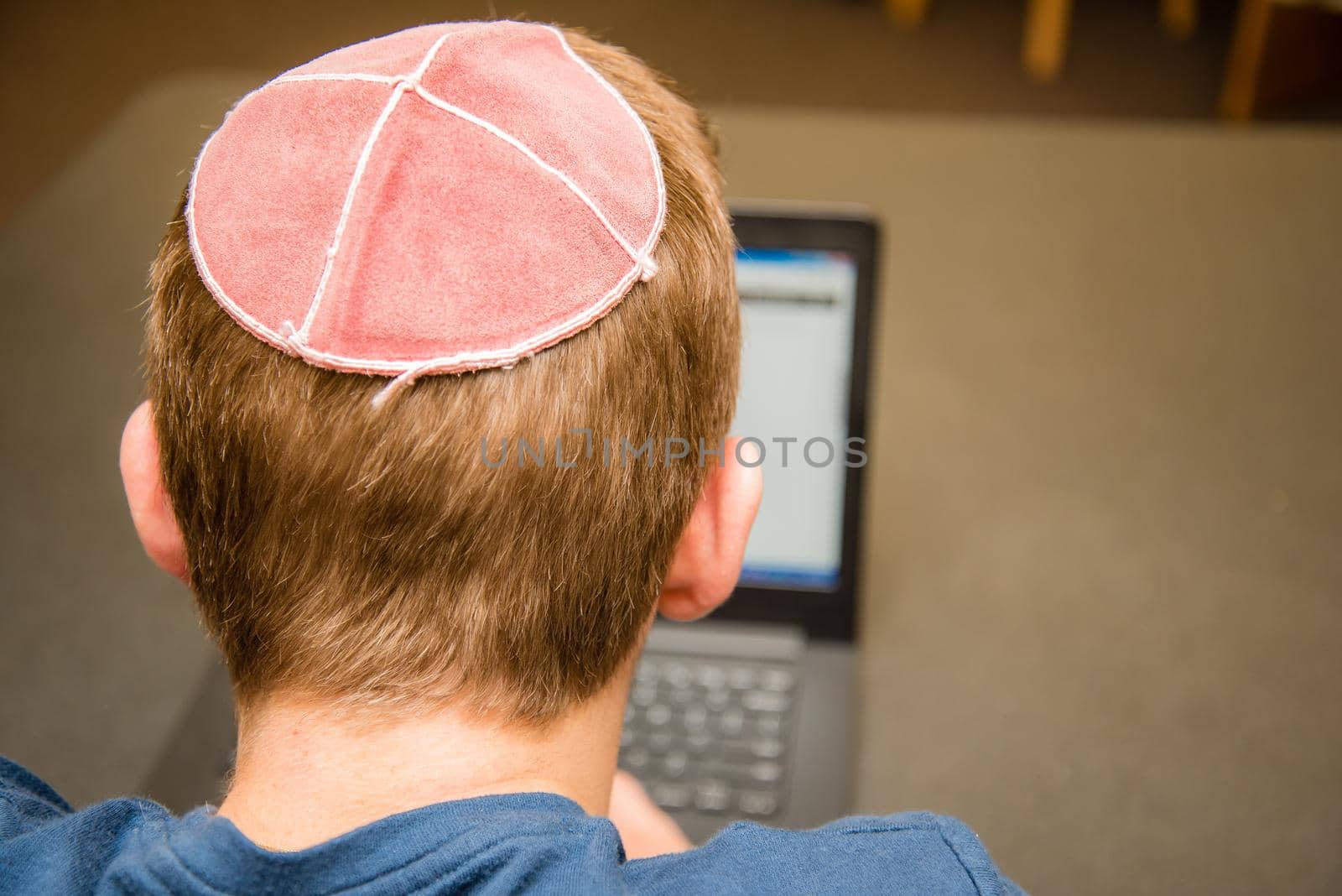 Young man wearing a yarmulke from the back doing work on a laptop in a library with colorful books on shelves.
