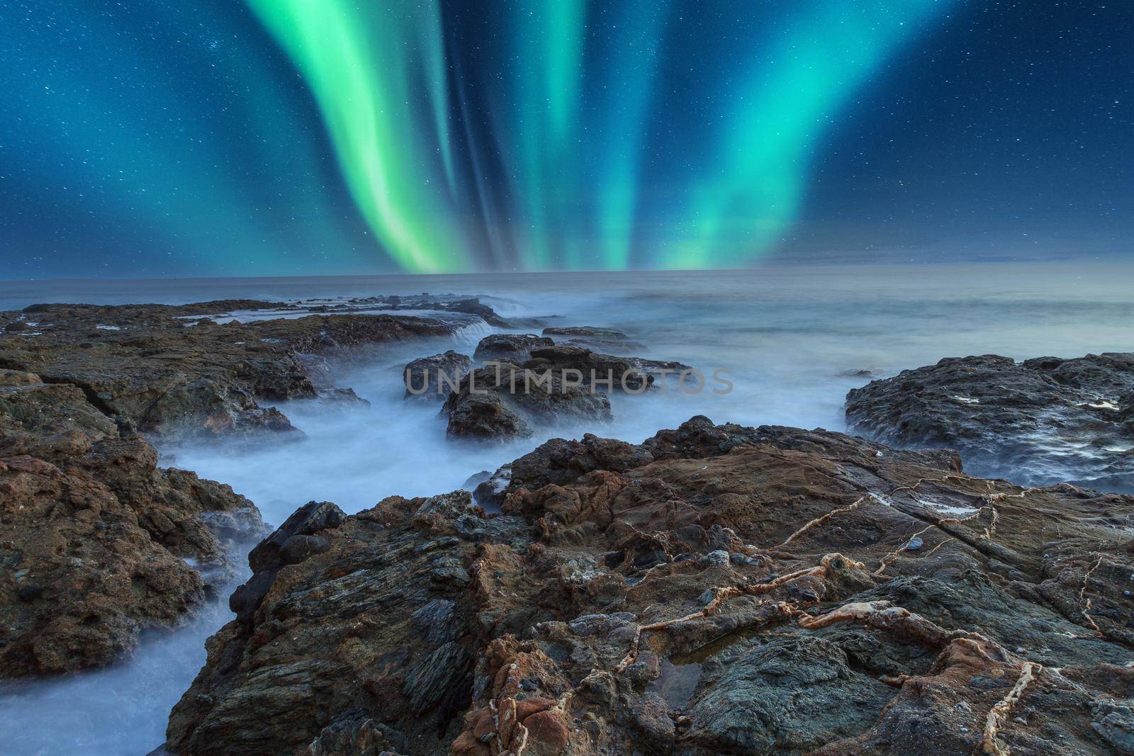 Green Aurora borealis shimmers over the ocean water as it cascades over rocks in Reykjavik, Iceland.