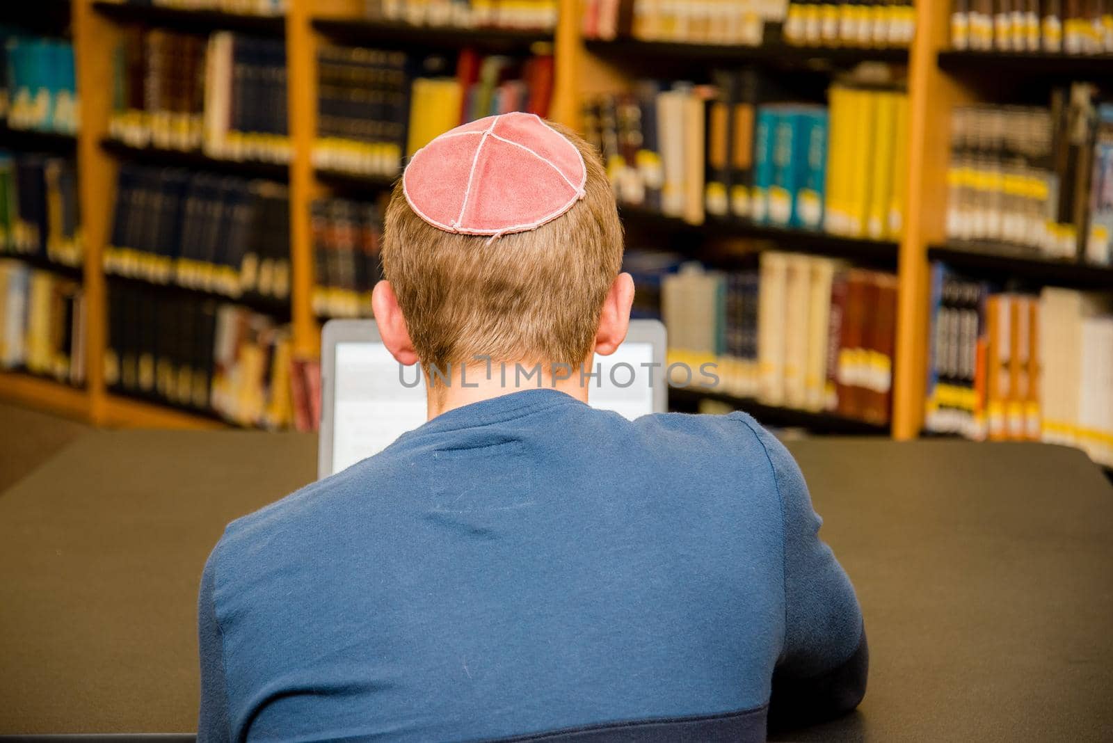 Young boy wearing a yarmulke from the back doing work on a laptop in a library with colorful books on shelves. by jyurinko