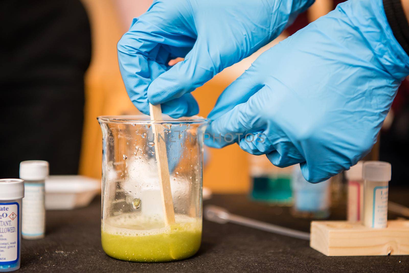 Up close view of scientist wearing blue latex gloves stirring green liquid in a chemistry glass.