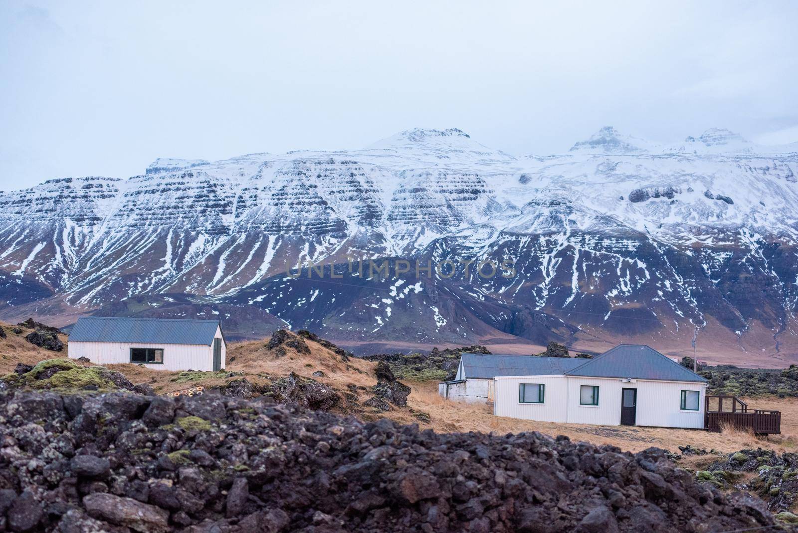 Landscape with Icelandic homes sitting in front of gorgeous snow capped mountains with layers of snow and dark rock. by jyurinko