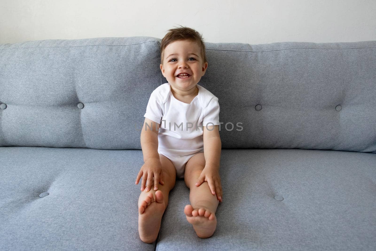 Funny happy healthy toddler one year old sitting at the sofa at home, pretty eyes, kids play and childhood side view