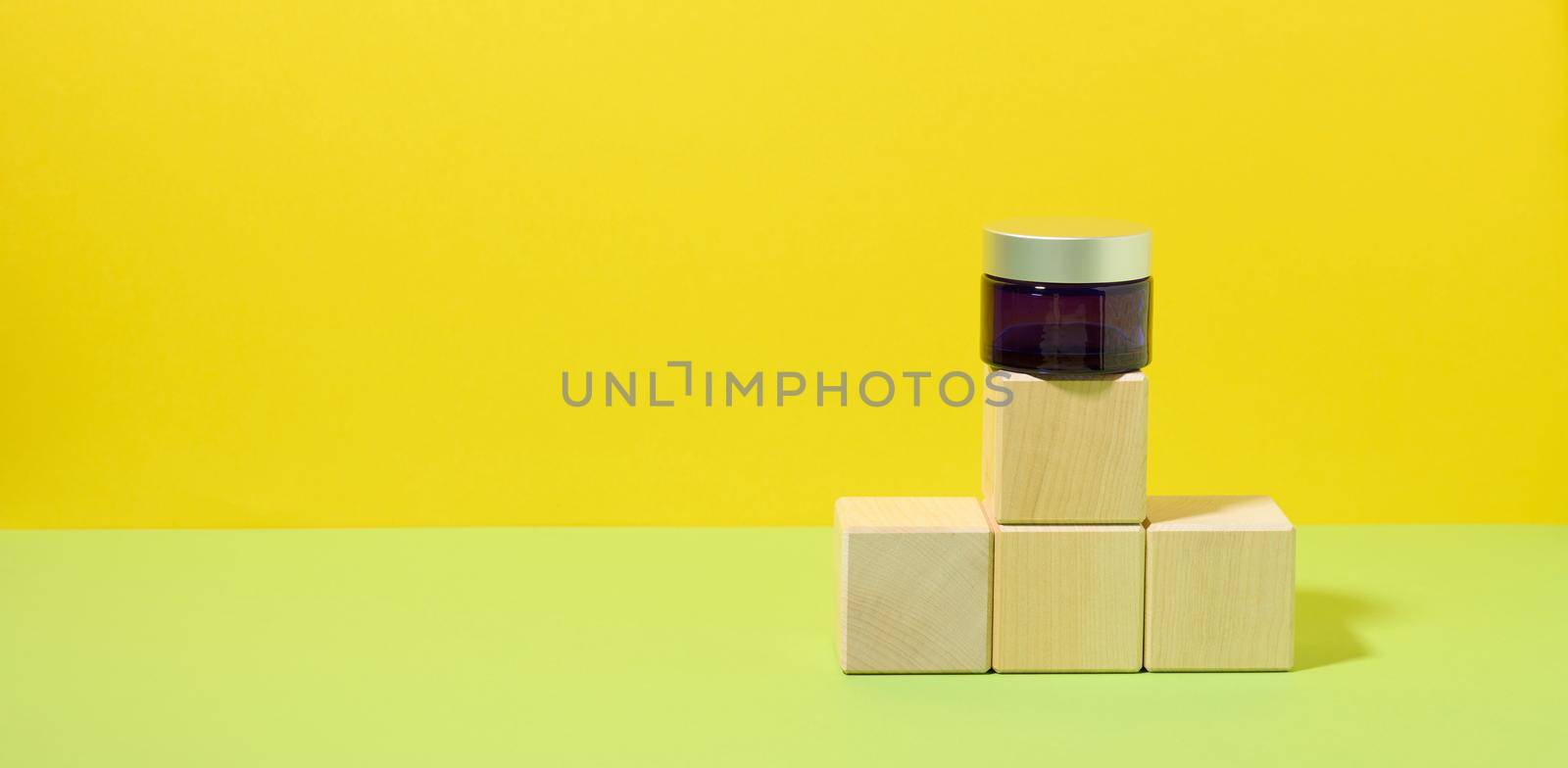 a blue glass jar with a gray lid stands on wooden cubes, yellow-green background. Cosmetic product display template by ndanko
